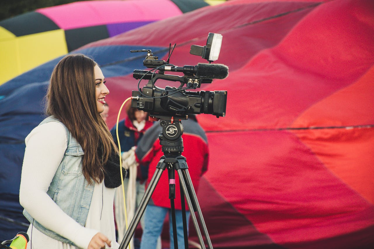 our-services-1 A female videographer operates a camera before a colorful hot air balloon backdrop.
