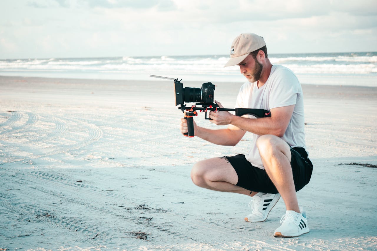 gallery-1 Young filmmaker capturing video on a sandy beach using handheld gear, enjoying the summer outdoors.