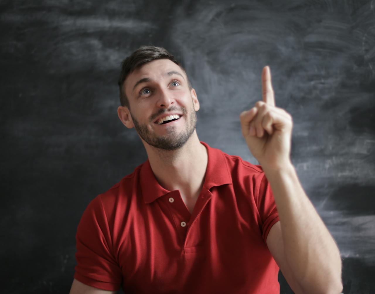 gallery-3 Happy young man in a red polo shirt smiling and pointing upwards in front of a blackboard.