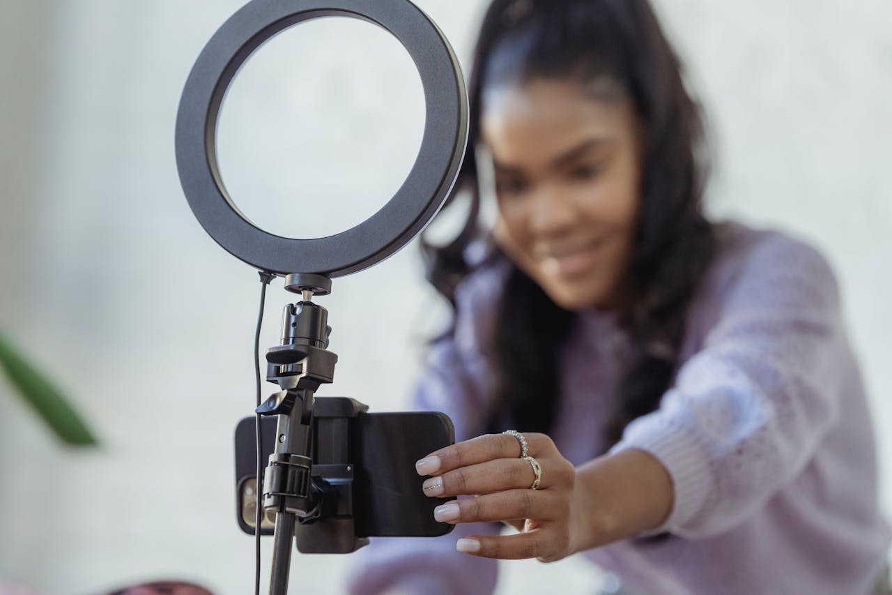 gallery-5 Cheerful young African American female blogger in stylish sweater smiling while setting up camera of smartphone attached to tripod with ring light before recording vlog